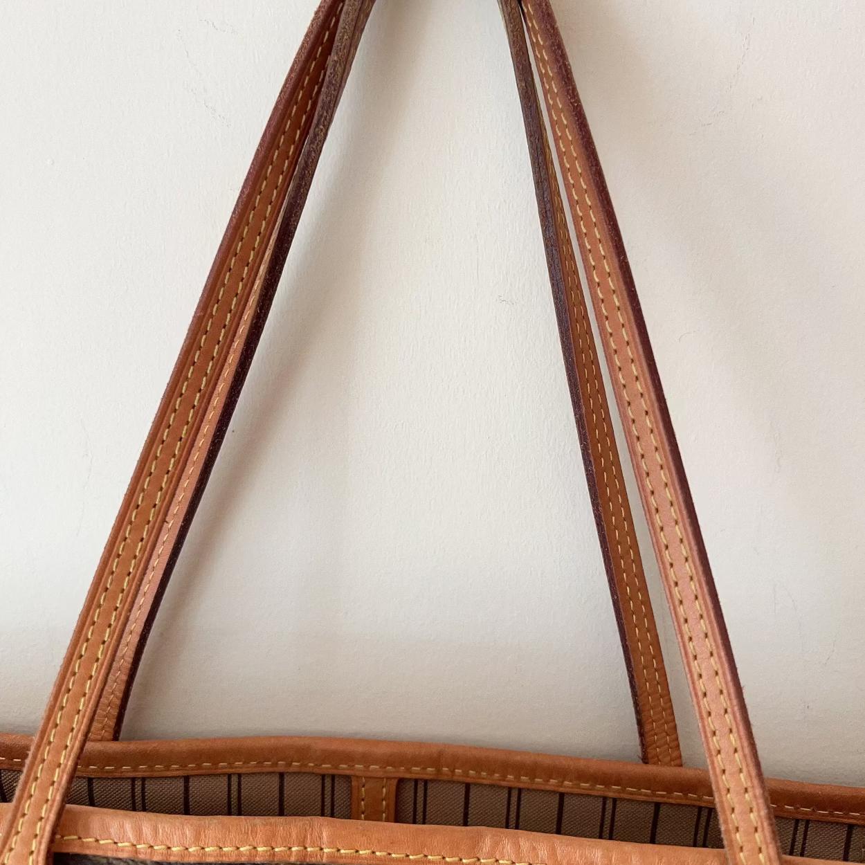 Close-up of a Louis Vuitton monogram tote bag with tan leather shoulder straps and a brown coated canvas body, hanging against a plain off-white wall.