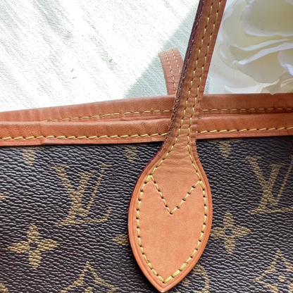 Close-up of a Louis Vuitton monogram bag in dark brown coated canvas with tan leather trim and a matching leather tab, photographed against a light fabric background.