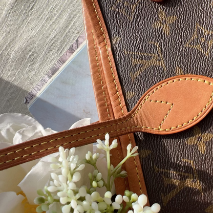 Close-up of a brown monogram handbag with tan leather trim and a matching leather strap tab, styled with white flowers on a light surface.