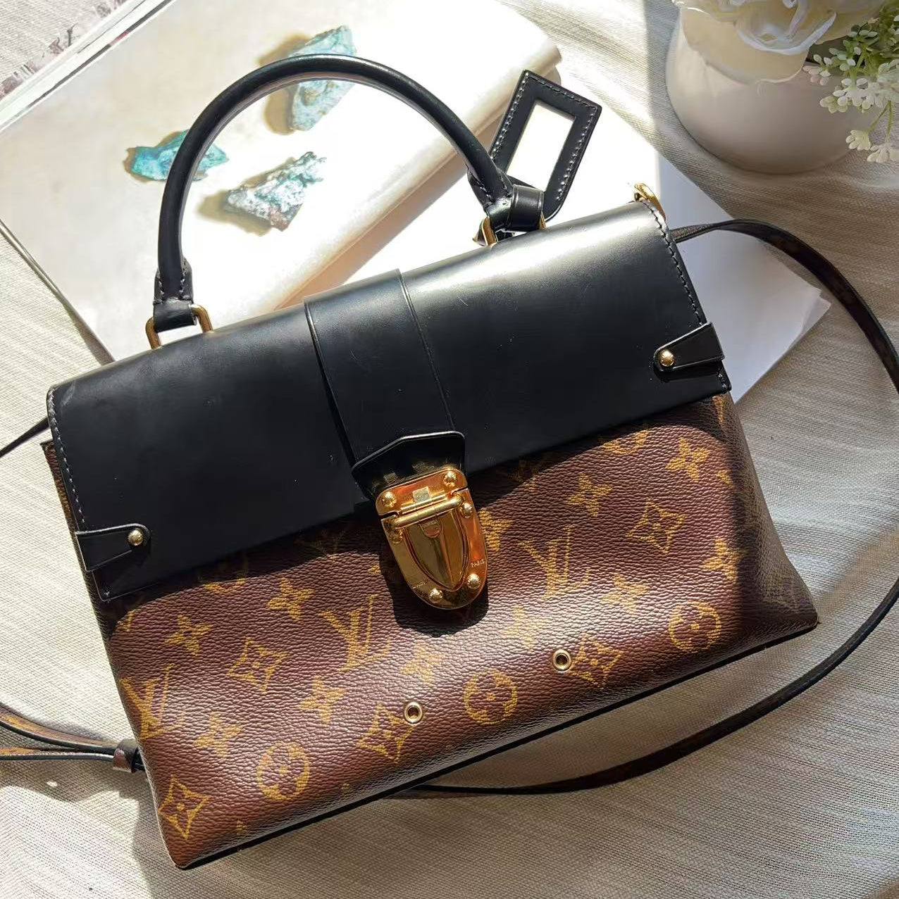 A Louis Vuitton monogram shoulder bag with a black leather top flap and handle, resting on a light-colored surface next to a book and a vase of flowers.