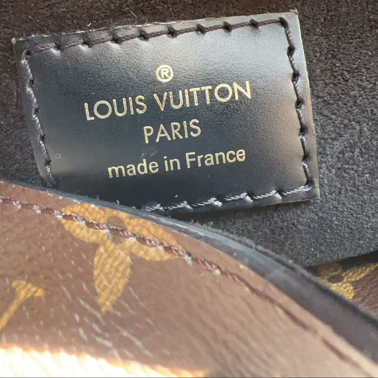 Close-up of a Louis Vuitton Paris interior label stitched onto a dark fabric surface, with part of a gold zipper and a finger visible at the top edge.