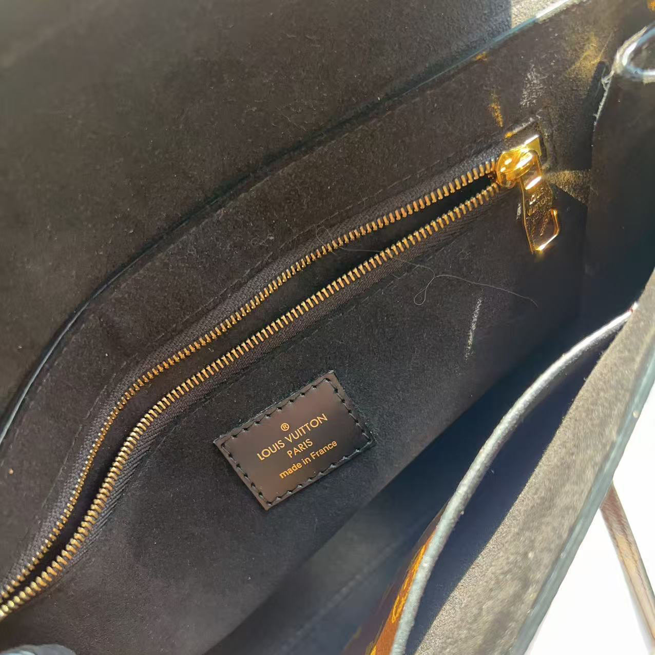 Close-up of the interior of a Louis Vuitton bag, showing a black fabric lining, a gold zipper, and a brown leather brand patch with gold lettering.
