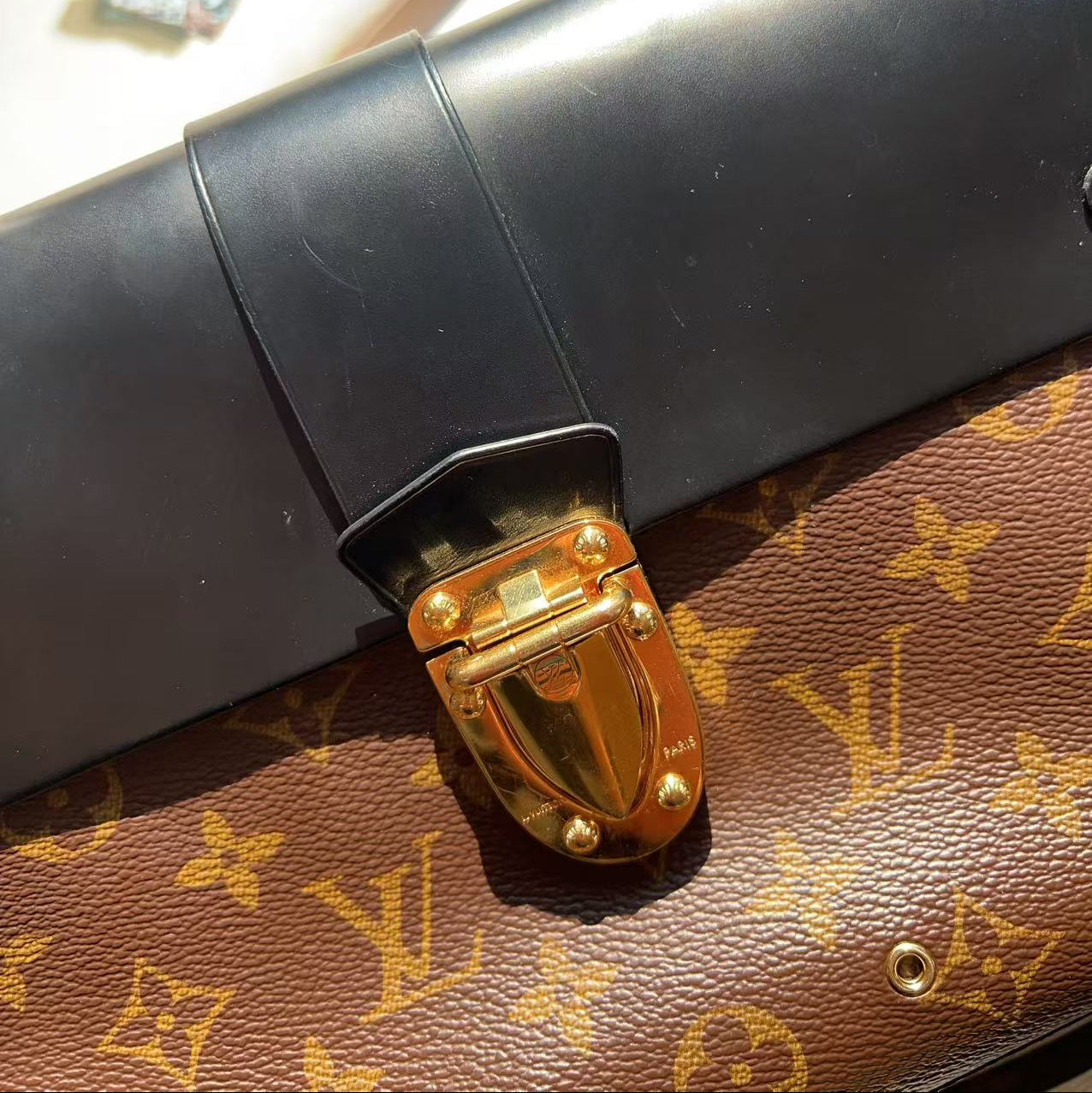 A close-up of a brown Louis Vuitton monogram canvas bag with a black leather flap and a prominent gold-tone metal clasp, resting on a light surface with crystals in the background.