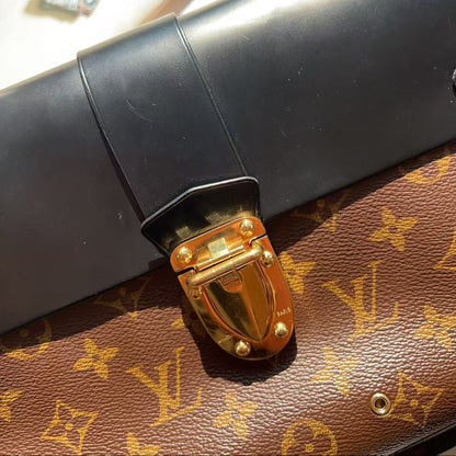 A close-up of a brown Louis Vuitton monogram canvas bag with a black leather flap and a prominent gold-tone metal clasp, resting on a light surface with crystals in the background.