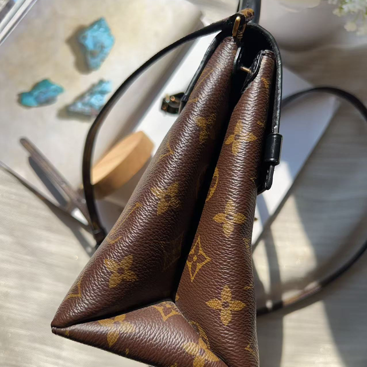 A close-up of a brown Louis Vuitton monogram handbag corner, showing the textured coated canvas with the classic yellow logo pattern and black leather trim.