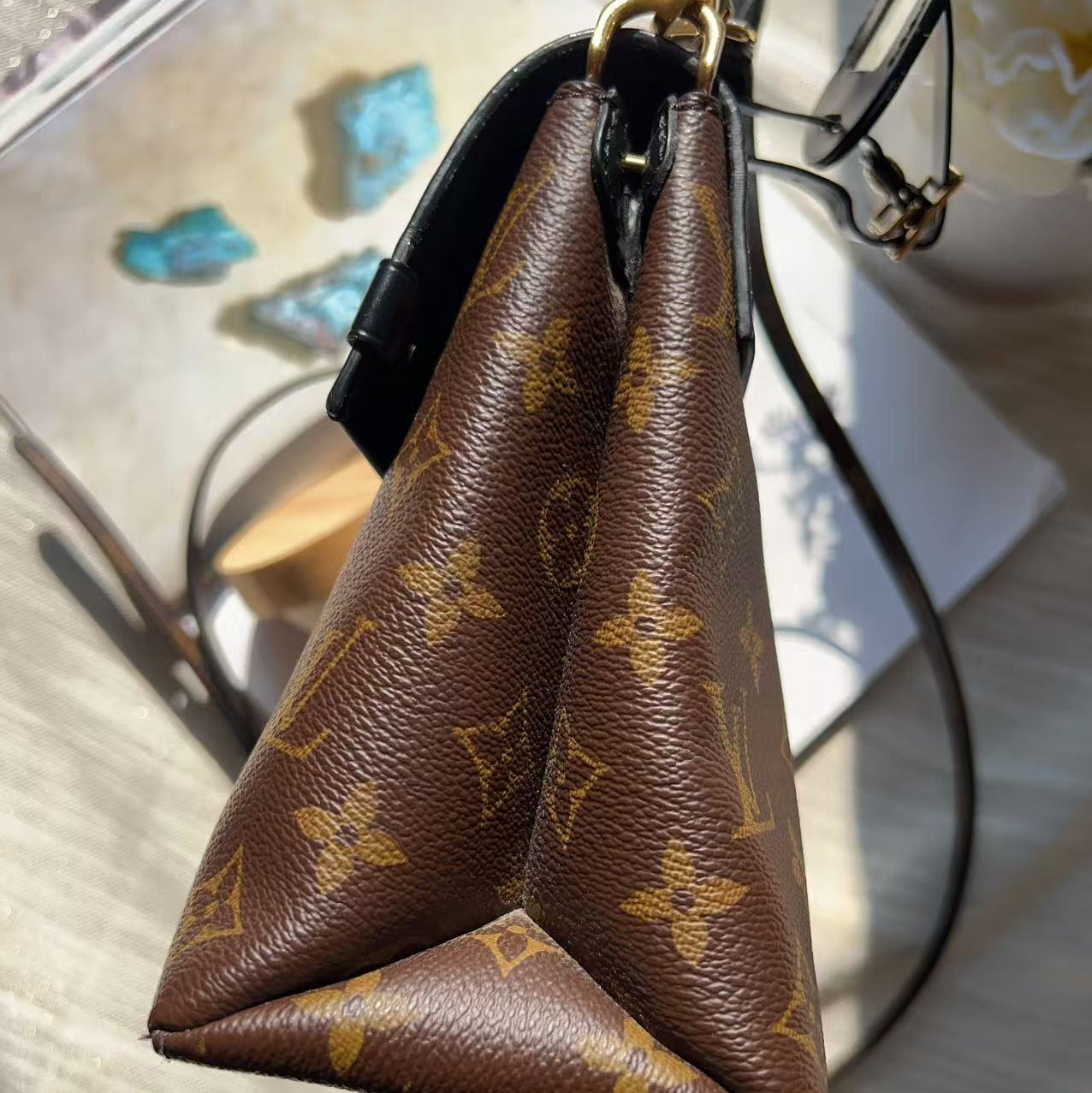 A close-up of a brown Louis Vuitton monogram handbag corner, showing the textured coated canvas with gold-tone monogram motifs and a black leather trim, resting on a light surface.