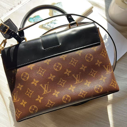 A Louis Vuitton monogram canvas handbag with a black leather top flap and handles, styled on a light-colored wooden surface with a book and flowers in the background.