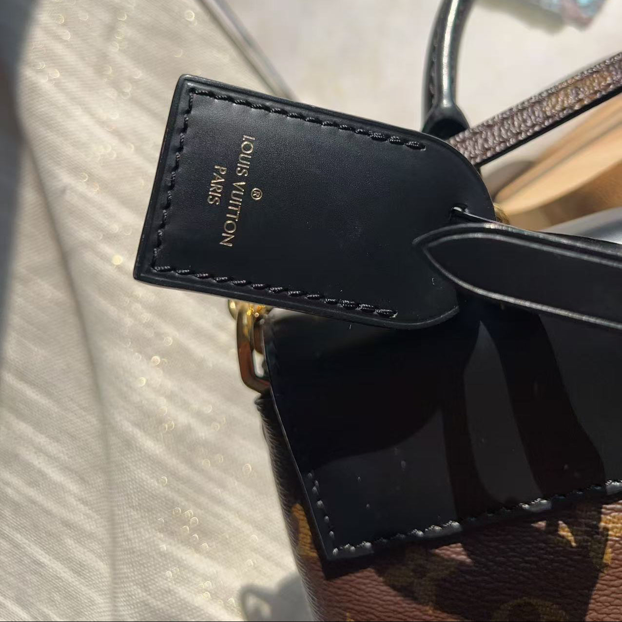 Close-up of a Louis Vuitton handbag tag in black leather with gold lettering, attached to a brown monogram bag with a glossy black handle.