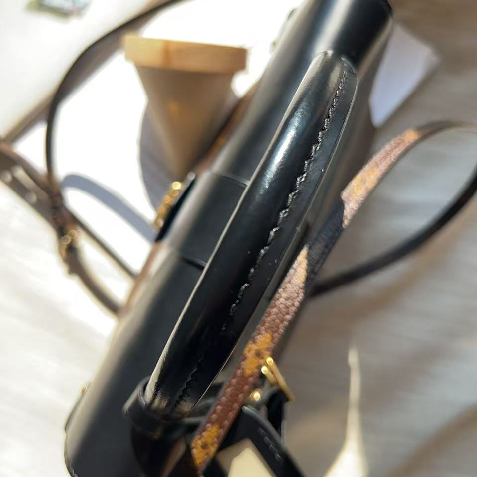 A close-up of a black leather shoulder bag with gold-tone hardware and a patterned brown strap, resting on a light fabric surface.
