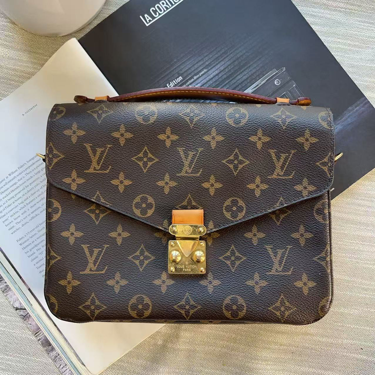 A Louis Vuitton monogram shoulder bag in brown coated canvas with gold-tone hardware, laid flat on top of books and a magazine on a light wood surface.