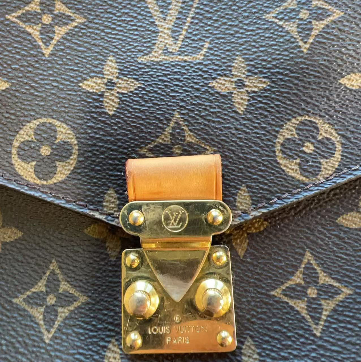 Close-up of a Louis Vuitton monogrammed bag, showing the dark blue coated canvas with beige monogram motifs and a gold-toned metal clasp with a light brown leather tab.