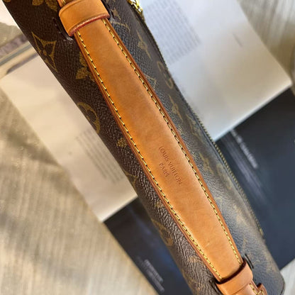 A close-up of a Louis Vuitton monogram shoulder bag in brown coated canvas with tan leather trim, resting on a light wood surface with books and a small plant in the background.