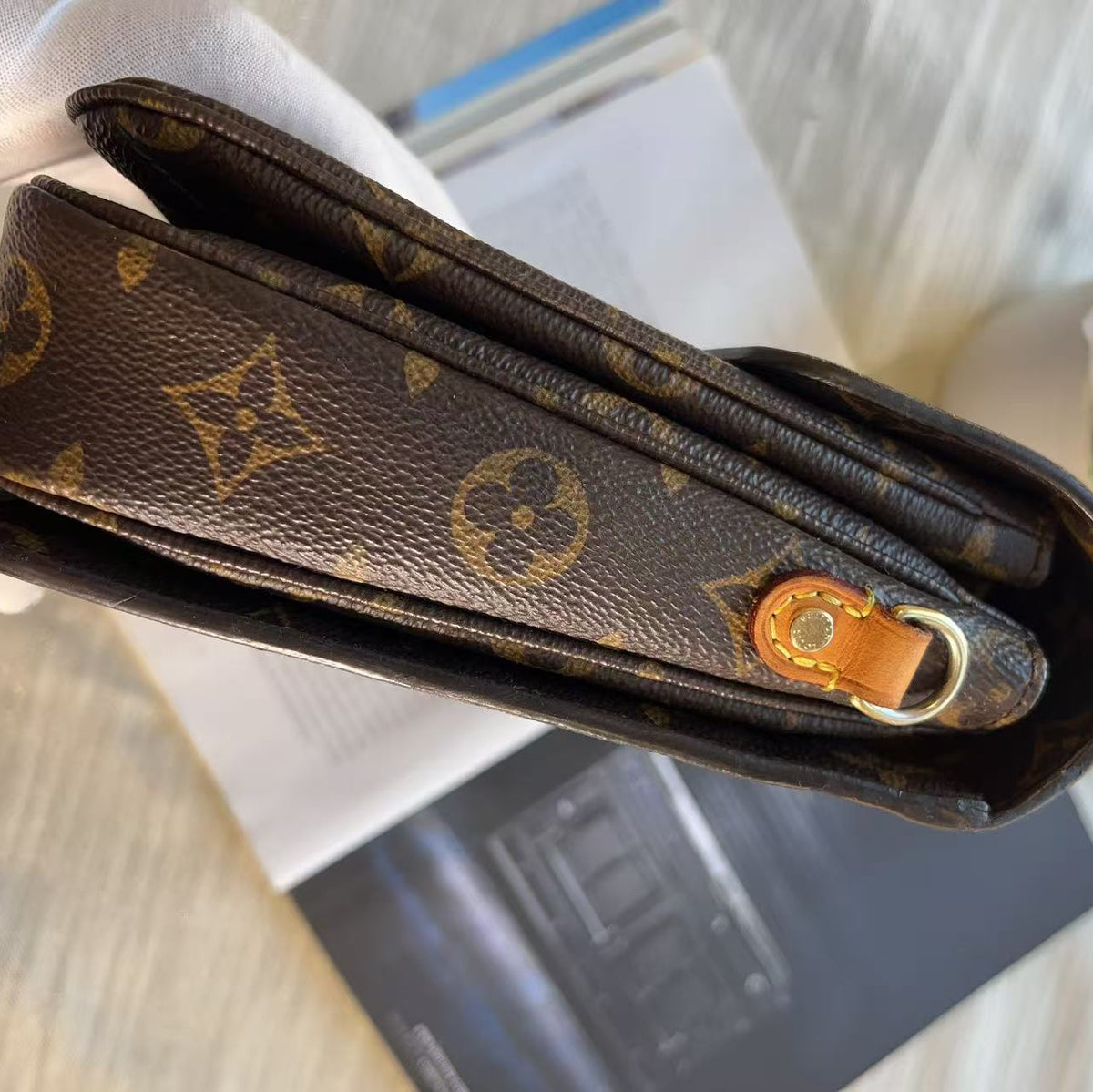 A close-up of a Louis Vuitton monogram shoulder strap held by a hand wearing a white glove, with a light wood surface and a book in the background.