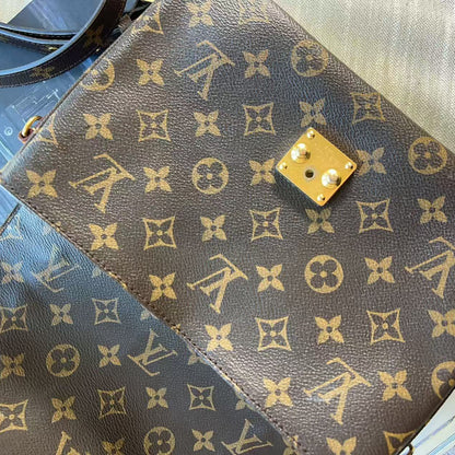 A close-up of a Louis Vuitton monogram canvas handbag in dark brown with gold-tone hardware, showing part of the flap and strap on a light fabric surface.