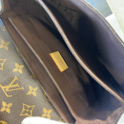 Close-up of the interior of a Louis Vuitton handbag, showing the dark brown fabric lining, a small gold brand plaque, and part of the classic brown and gold monogram canvas. A white gloved hand is holding the bag open.