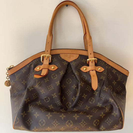 A Louis Vuitton monogram canvas shoulder bag with tan leather handles and trim, hanging against a plain off-white wall.