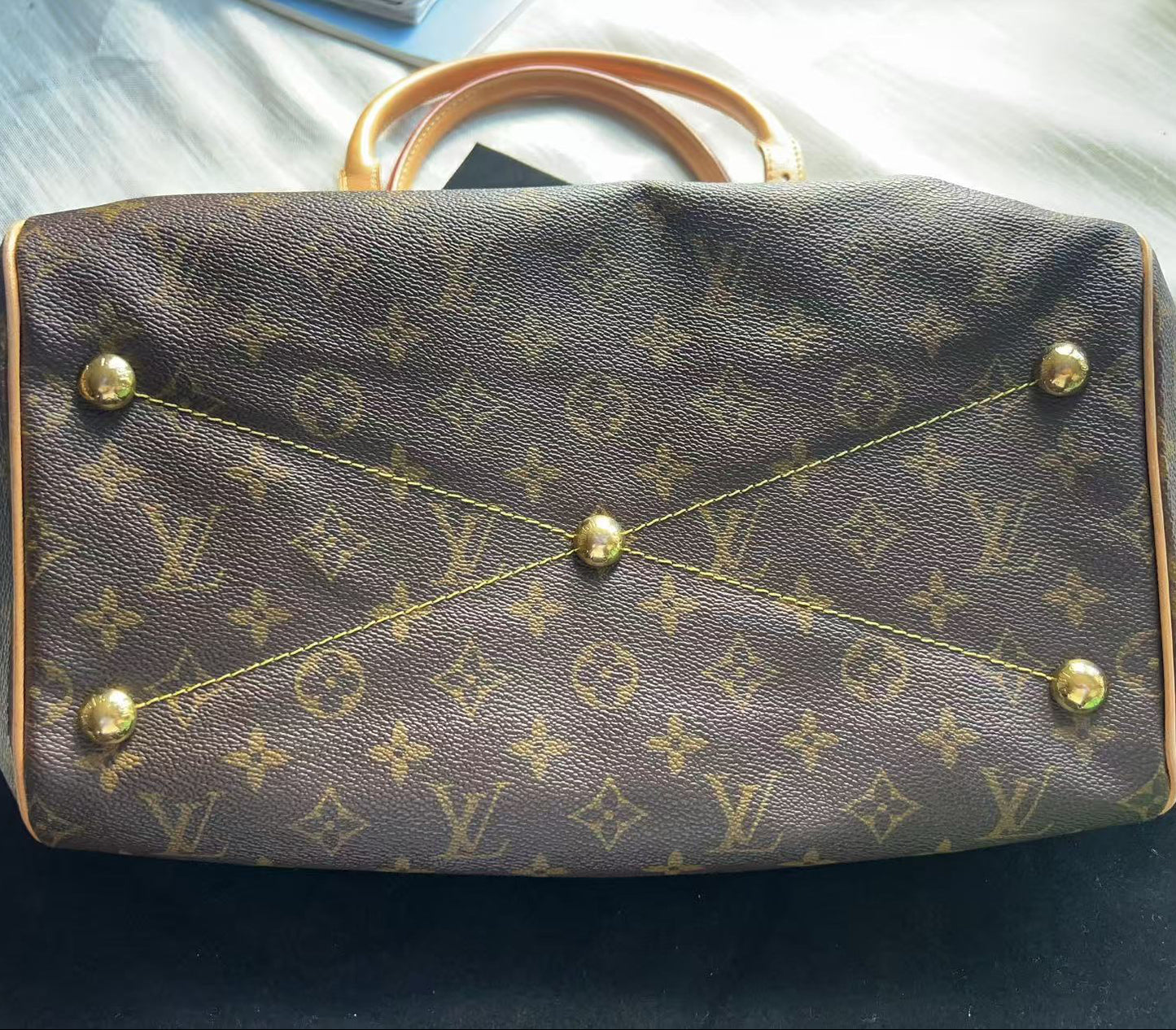 A Louis Vuitton monogram canvas handbag shown from the front, featuring the classic brown and tan pattern with gold-tone hardware and a light tan leather handle, resting on a dark surface with a light-colored fabric background.