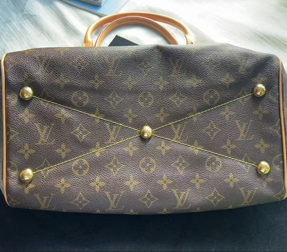 A Louis Vuitton monogram canvas handbag shown from the front, featuring the classic brown and tan pattern with gold-tone hardware and a light tan leather handle, resting on a dark surface with a light-colored fabric background.