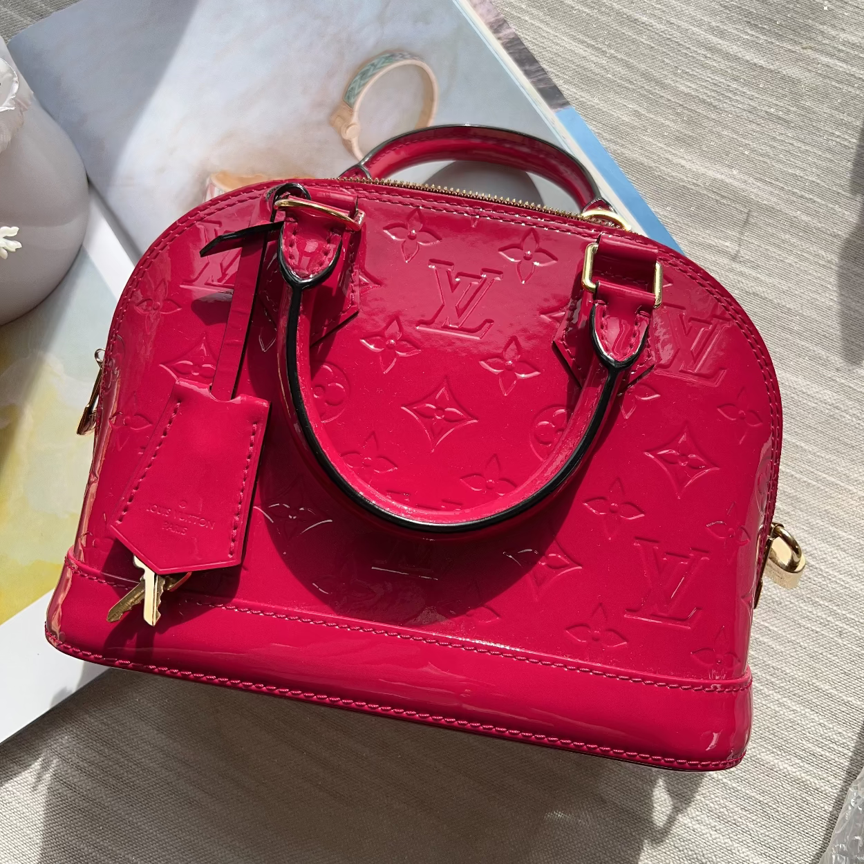 A bright pink Louis Vuitton monogram handbag with gold-tone hardware, resting on a light-colored wooden surface next to a magazine.