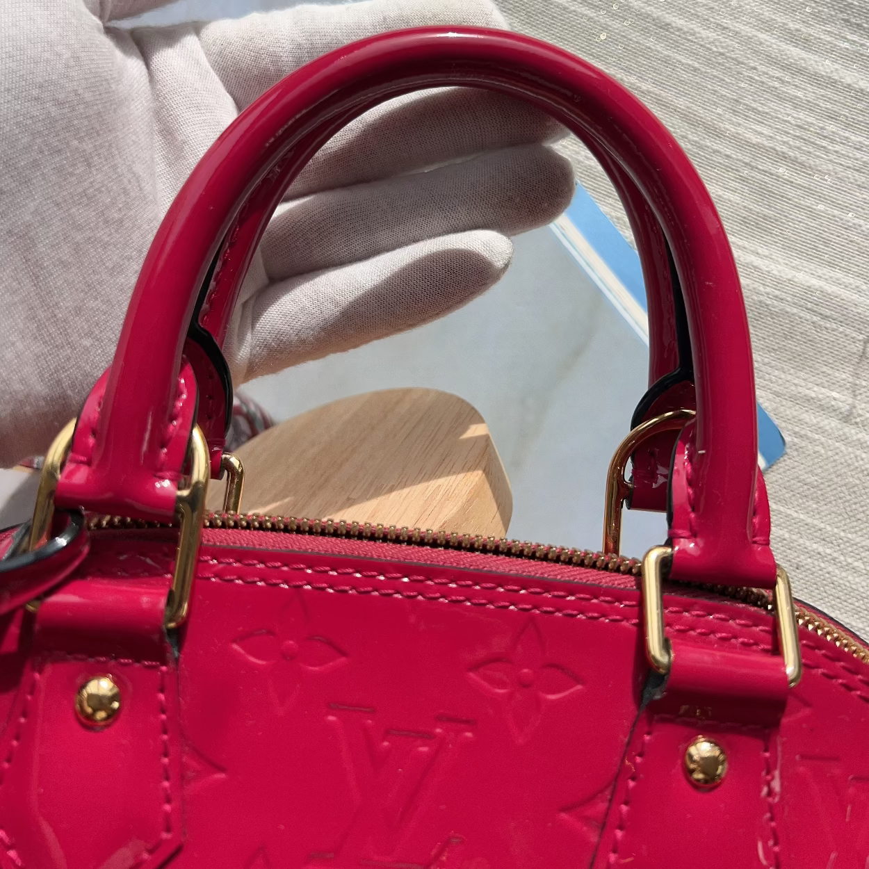A close-up of a bright pink Louis Vuitton handbag with gold-tone hardware, held by a hand wearing a white glove. The embossed LV logo is visible on the front, and the bag is shown against a light gray textured background.