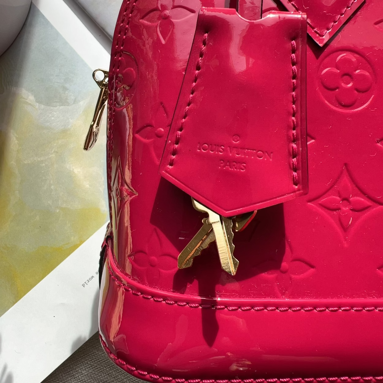 Close-up of a Louis Vuitton patent leather handbag in bright pink with embossed monogram pattern and gold-tone hardware, including a hanging keychain tag.