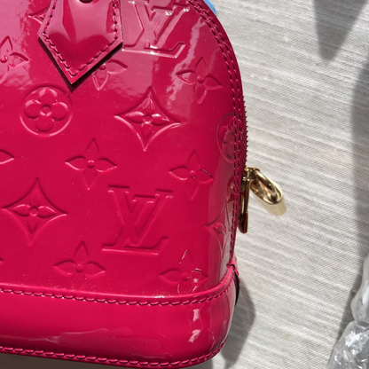 Close-up of a glossy pink Louis Vuitton monogram handbag with gold-tone hardware, photographed on a light gray textured surface.