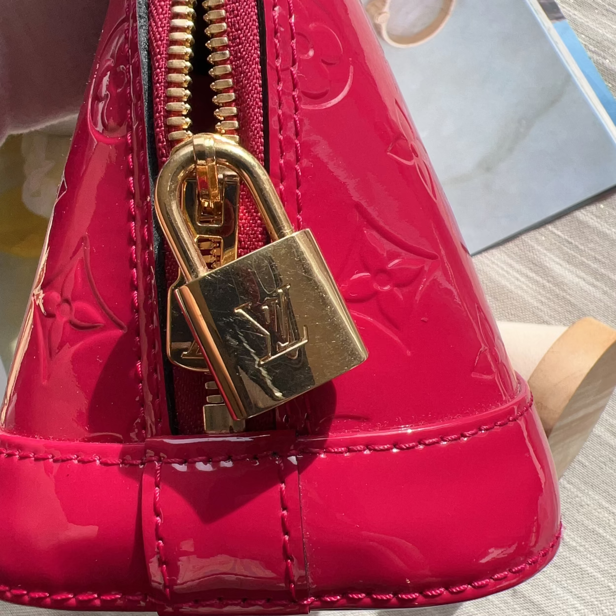 A close-up of a bright pink patent leather handbag with an embossed monogram pattern, featuring a gold-tone padlock zipper pull and gold zipper track.