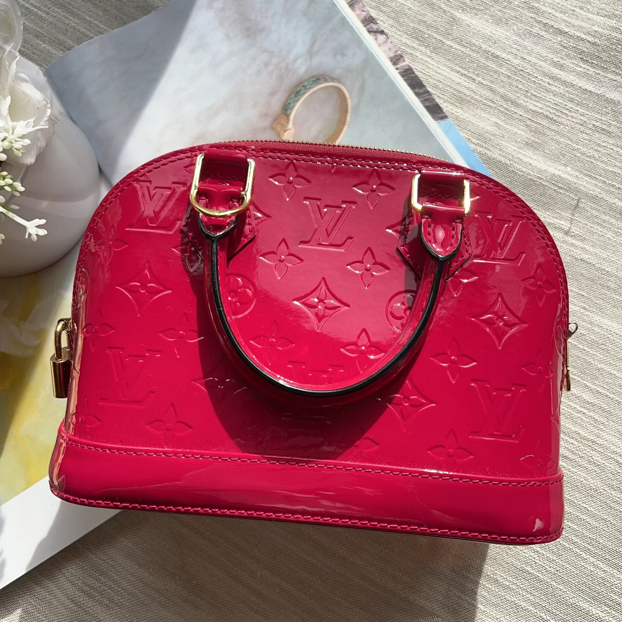 A red Louis Vuitton monogram coated canvas handbag with gold-tone hardware, displayed on a light wood surface next to a white ceramic vase.
