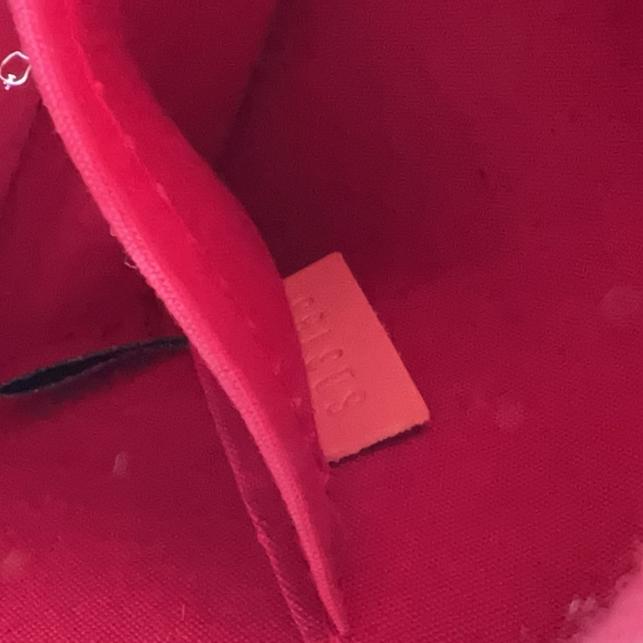 Close-up of the interior lining of a bright pink or magenta bag, showing a small rectangular brand tag with embossed lettering and a black tag attached near the edge.