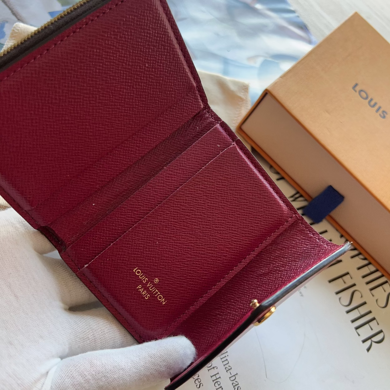 Red wallet with gold accents held by a gloved hand, with a Louis Vuitton box in the background.