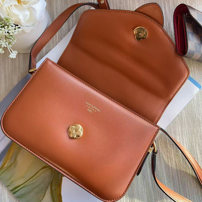 Brown leather handbag with a gold clasp on a wooden surface with flowers and a book.
