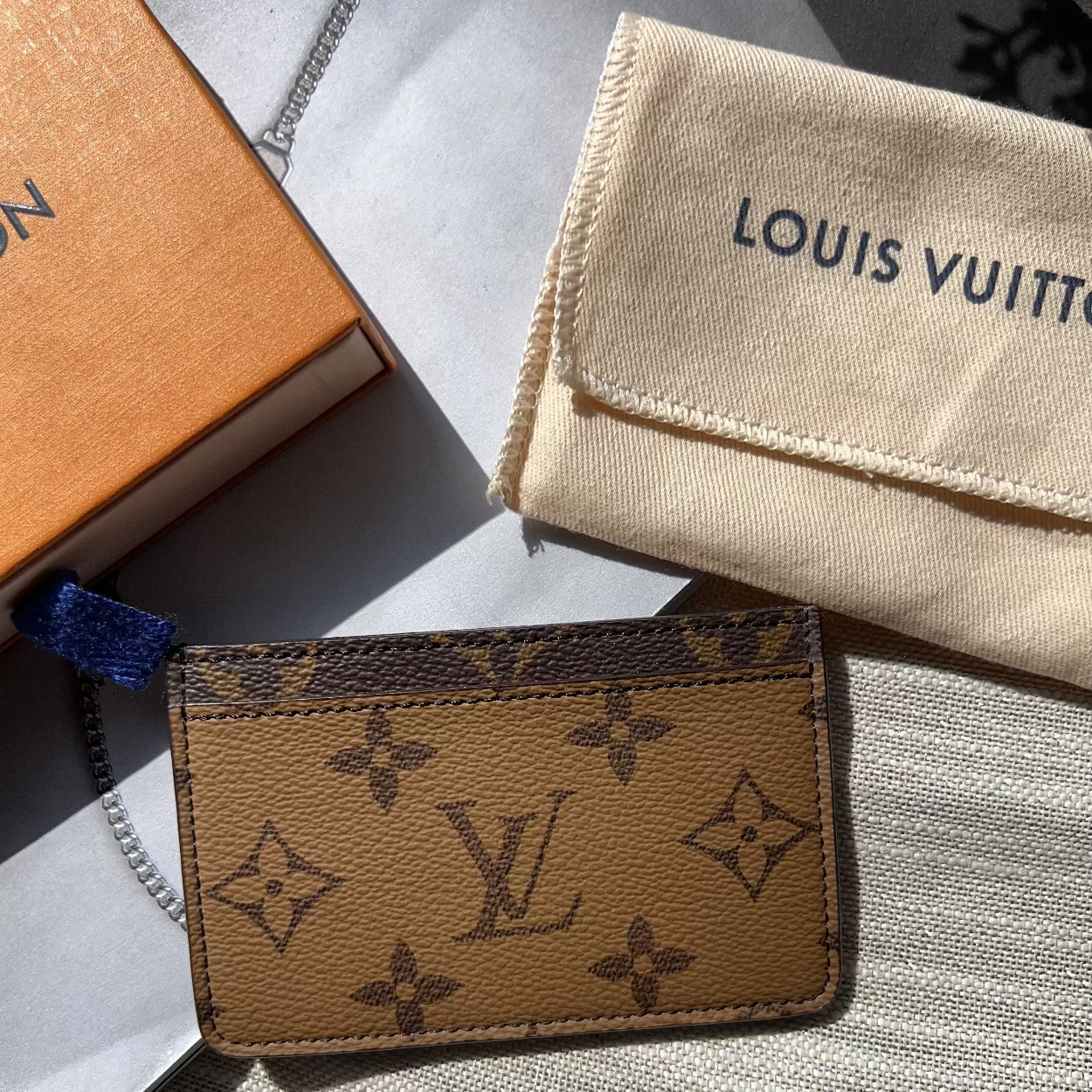 Louis Vuitton cardholder with monogram pattern on a light gray surface, with a branded cloth and box in the background.