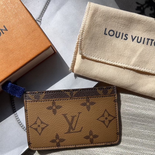 Louis Vuitton cardholder with monogram pattern on a light gray surface, with a branded cloth and box in the background.