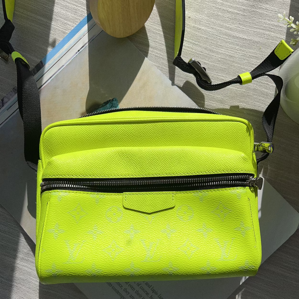 A bright neon yellow-green crossbody bag with a subtle monogram pattern, black zippers, and black straps, resting on a light-colored wooden surface with papers underneath.