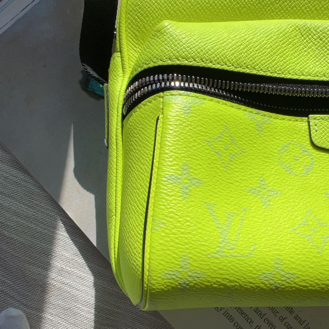 A close-up of a bright neon yellow-green Louis Vuitton monogram bag, showing the textured canvas material and a black zipper, resting on a light-colored surface.