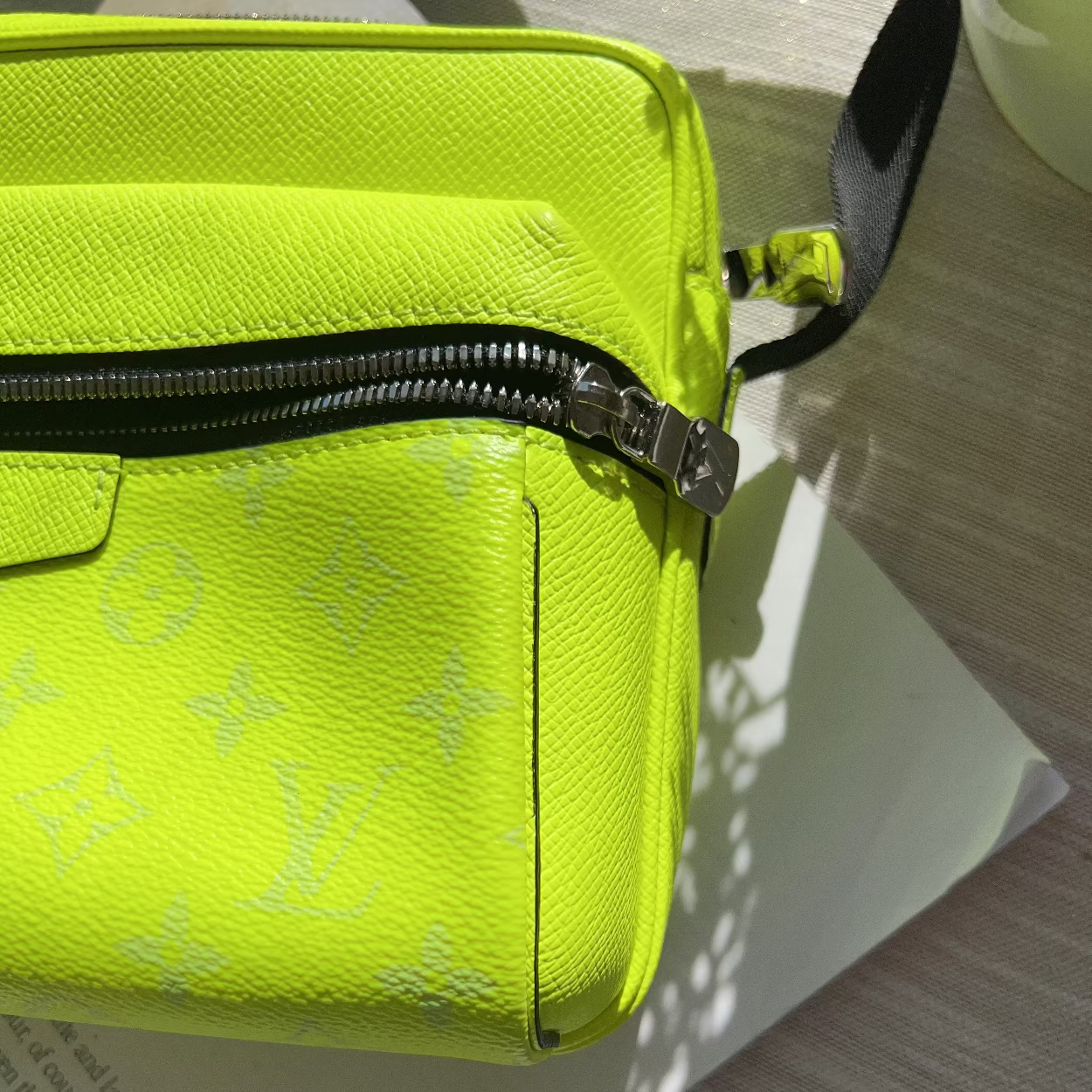 A close-up of a bright neon yellow-green Louis Vuitton crossbody bag in textured monogram canvas, resting on a white sheet of paper over a light wood surface.