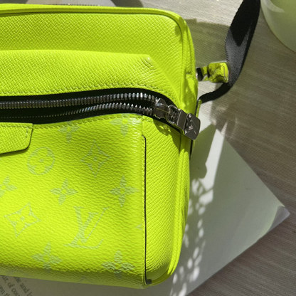 A close-up of a bright neon yellow-green Louis Vuitton crossbody bag in textured monogram canvas, resting on a white sheet of paper over a light wood surface.