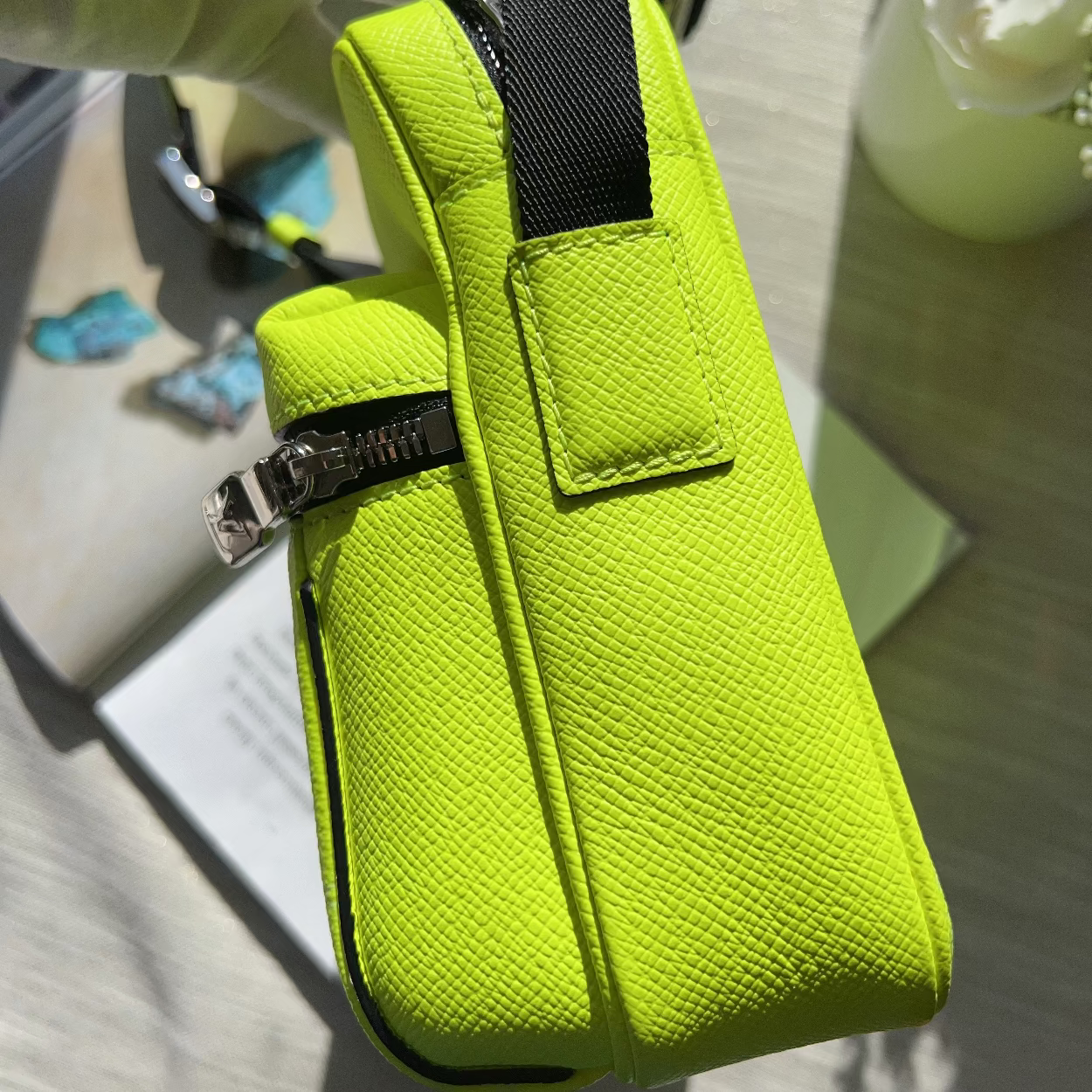 A bright neon yellow-green textured leather pouch bag with a black webbed strap and silver zipper, held by a hand in a light green glove.