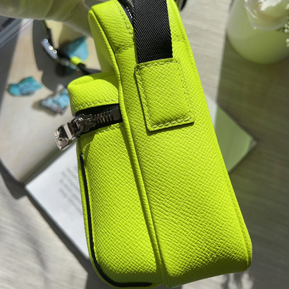 A bright neon yellow-green textured leather pouch bag with a black webbed strap and silver zipper, held by a hand in a light green glove.