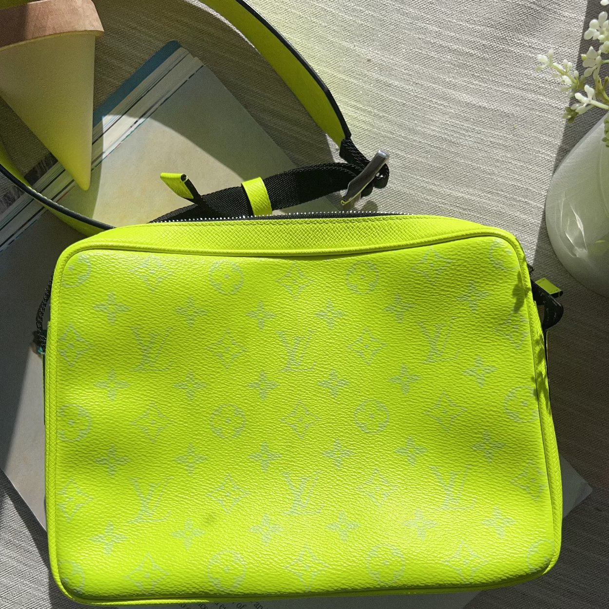 A bright neon yellow-green crossbody bag with a subtle repeating monogram pattern, resting on a light-colored wooden surface with some papers and a vase in the background.