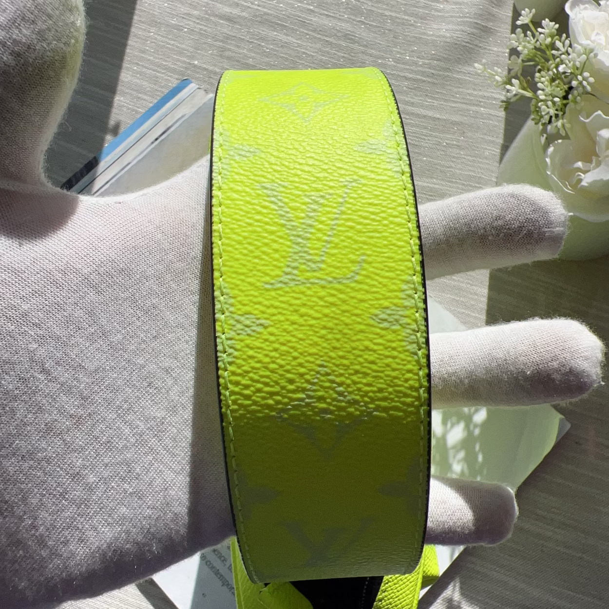 A close-up of a bright neon yellow-green Louis Vuitton monogram canvas belt being held by a white gloved hand, with a light-colored wooden surface and small flowers in the background.