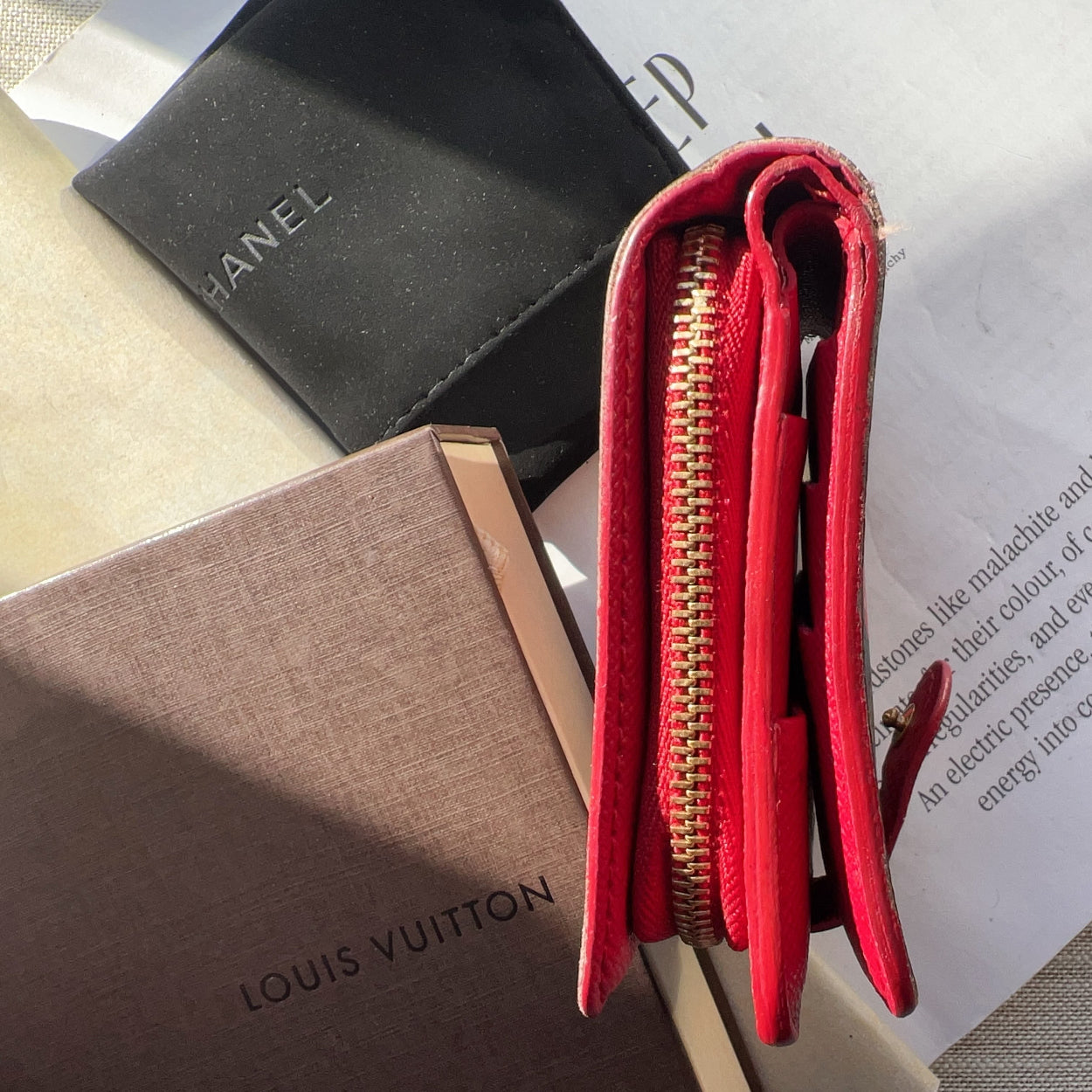 Red wallet with gold zipper on a surface with a Chanel and Louis Vuitton box in the background.