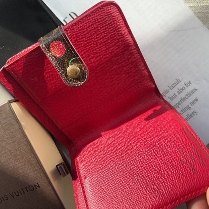 Red wallet with gold clasp held by a hand, with a blurred background featuring a Louis Vuitton bag.