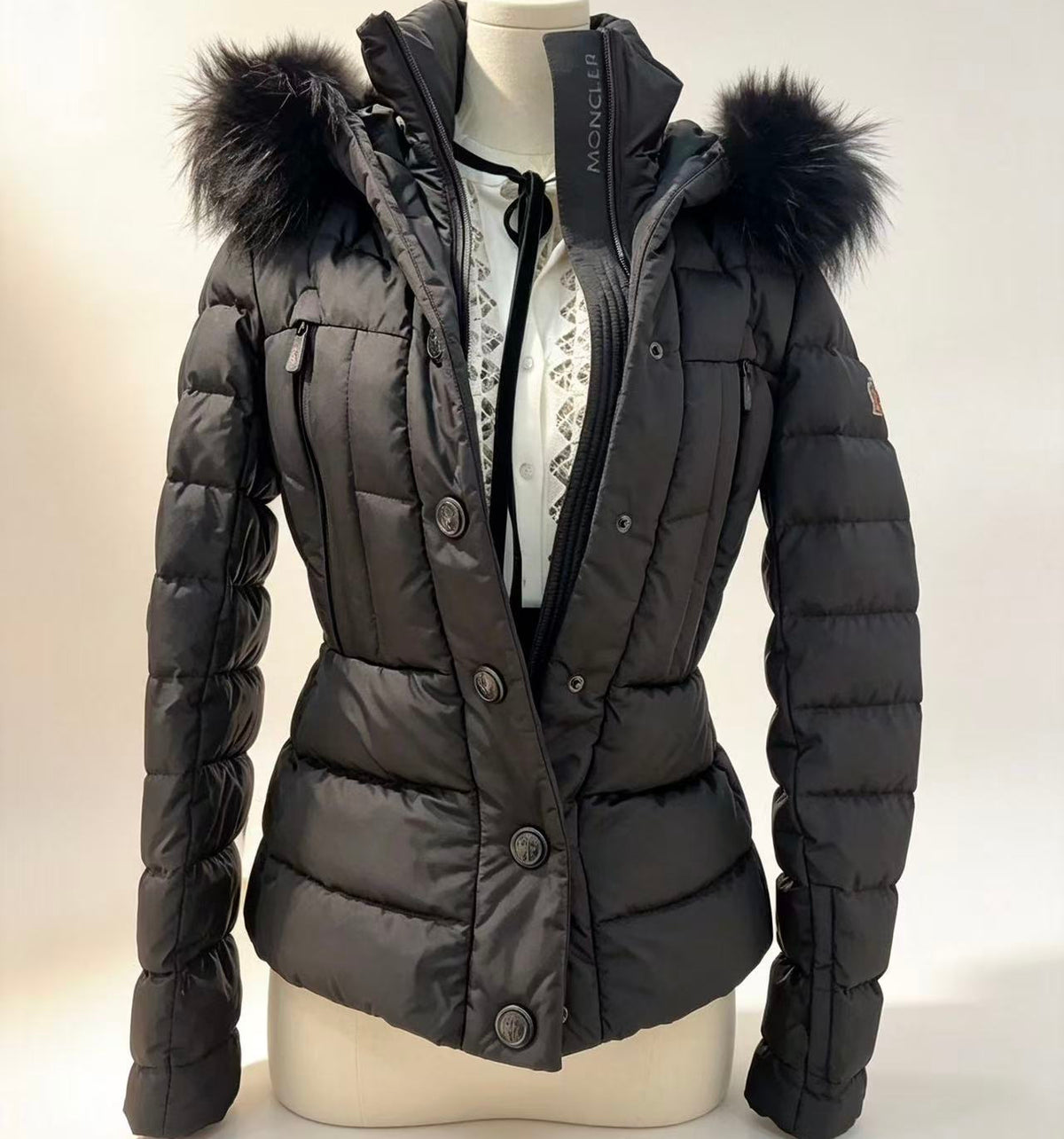 A black Moncler down puffer jacket with a black fur-trimmed hood, displayed on a cream mannequin torso against a light beige background.