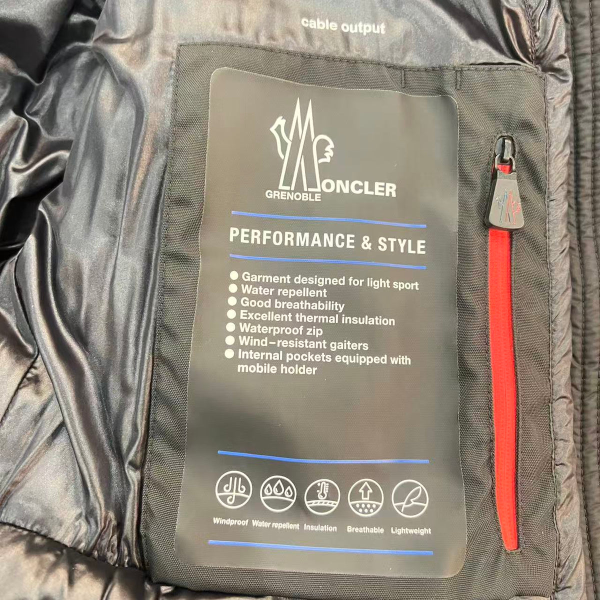 Close-up of the inside of a dark gray waterproof jacket showing a large gray label with the Moncler brand name and product features, next to a red zipper pull.