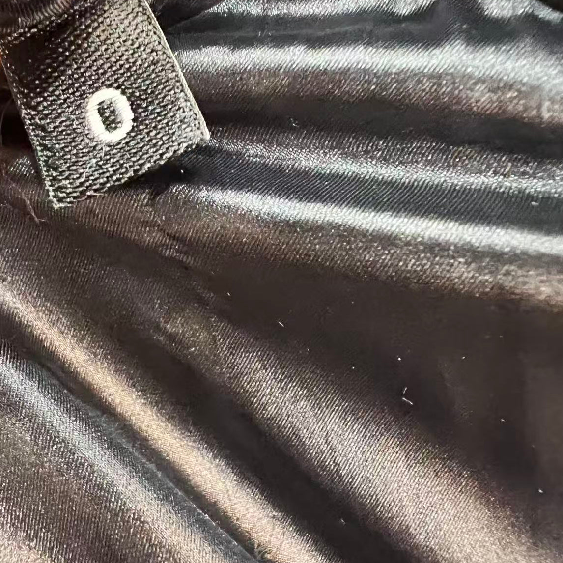 Close-up of a black satin-like garment interior with a small fabric tag attached, held by fingers. The tag has a white circular emblem, and the surrounding material is shiny black.