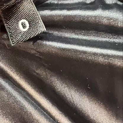 Close-up of a black satin-like garment interior with a small fabric tag attached, held by fingers. The tag has a white circular emblem, and the surrounding material is shiny black.