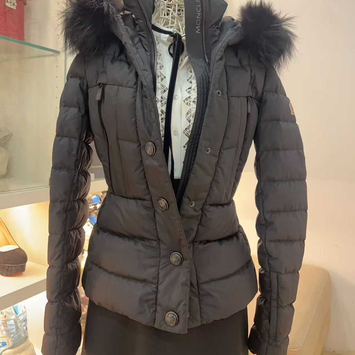 A dark gray Moncler puffer jacket with a black fur-trimmed hood, displayed on a dress form mannequin in a retail store setting.