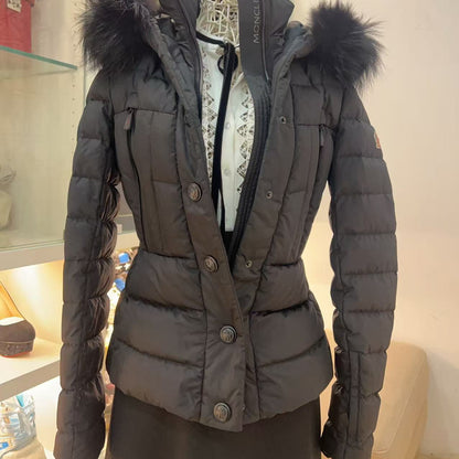 A dark gray Moncler puffer jacket with a black fur-trimmed hood, displayed on a dress form mannequin in a retail store setting.