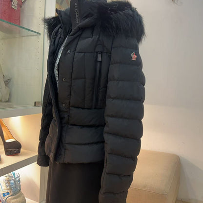 A black puffer jacket with a dark faux-fur hood trim displayed on a mannequin torso inside a retail store. The jacket features horizontal quilting, a front zipper, and a small embroidered logo on the left sleeve.